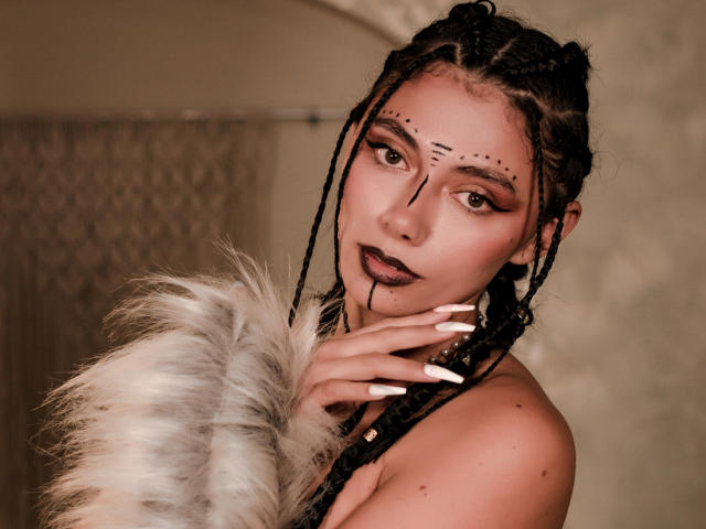 Anna Mooree mit Zöpfen, dunklem Lippenstift und Gesichtspunkten, die ein flauschiges, weißes Kleidungsstück hält und dabei ihre langen, weißen Nägel zur Schau stellt, auf einem Foto mit weichen, warmen Farbtönen..