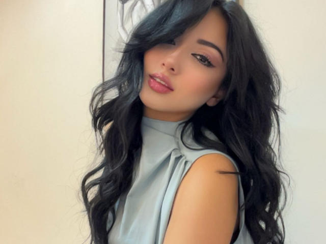 LillyTaylor with long black wavy hair, light gray sleeveless top, pink lipstick, subtle eye makeup, and a cute, slightly cocky expression. Her ass is barely visible.