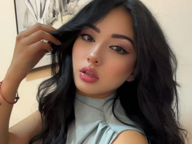 LillyTaylor has long black hair, pink glossy lips, and is wearing a light gray sleeveless top. She's looking at the camera with a slight hand in her hair.