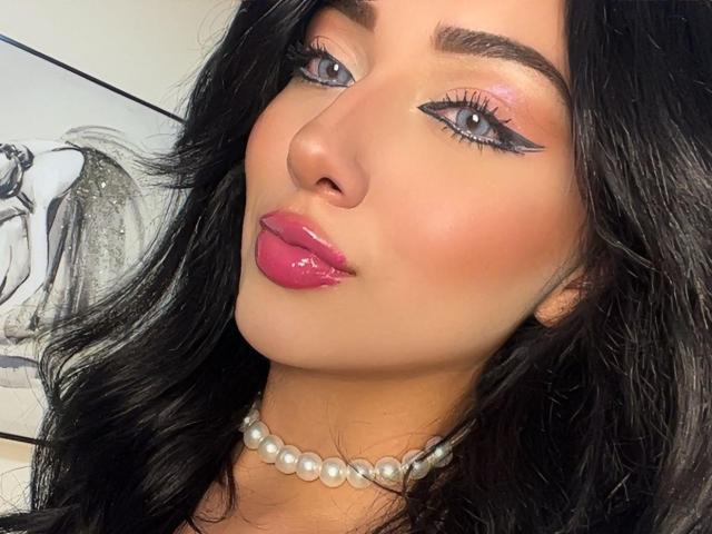 LillyTaylor has shiny black hair, pink glossy lips, blue eyes with black eyeliner, and a pearl necklace. Her face is slightly tilted, showing a cute, sexy expression.