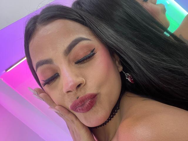 Amber, con los ojos cerrados, sombra de ojos rosa anaranjada, delineador de ojos grueso, labios rojos brillantes, piercing en la nariz, collar negro, cabello negro largo, aretes rojos y uñas pintadas, haciendo un puchero en un selfie..