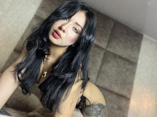 LillyTaylor with long black hair, blue eyes, red lips, and a gold necklace, leaning forward in black lingerie, showing her tattooed right thigh and small, perky tits.