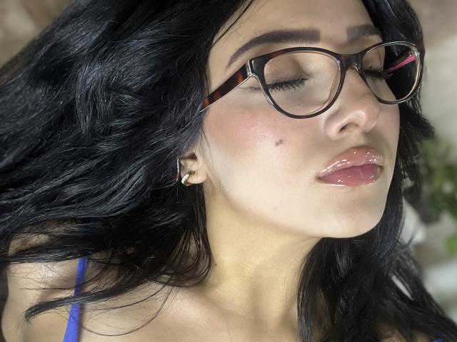 LillyTaylor with black wavy hair, tortoiseshell glasses, glossy pink lips, slight blemish on right cheek, blue spaghetti strap top, eyes closed, looking relaxed and sexy.