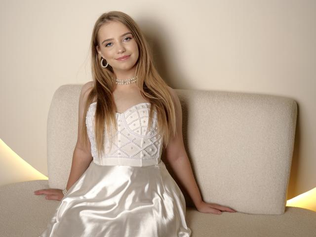 LunaBellaHottiest with long blonde hair, wearing a white lace bodice dress and satin skirt, sitting on a beige sofa, smiling, pearl necklace, and earrings.