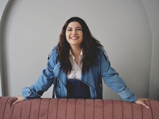 CrimsonDusk, smiling with dark wavy hair, wearing a blue denim jacket and white shirt, leaning on a pink upholstered bench, hands on top, tits visible under shirt.