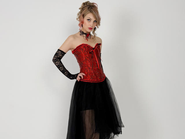 LindaAndersoni with red lace corset, black lace gloves, and black tutu, standing with hands on hips, blonde curly hair, red lipstick, pale skin, looking at camera.
