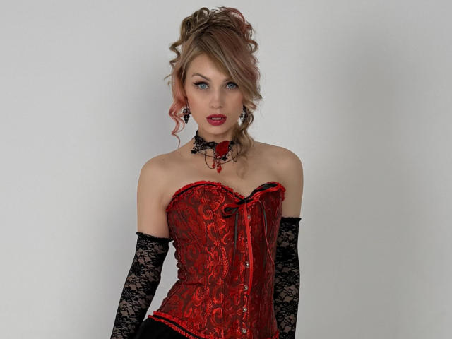LindaAndersoni has red lace-up corset, black lace gloves, pink curly hair, red lipstick, and black choker with red heart, showing her tits and looking sexy.