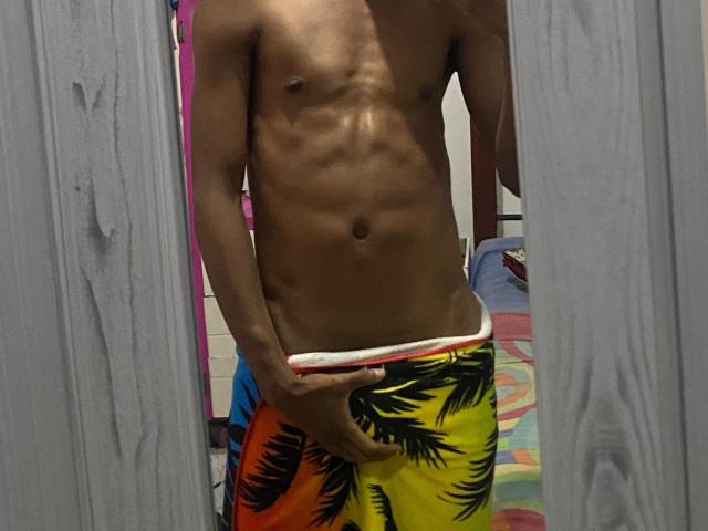 BrownyX's slim, toned, dark brown body with visible abs and small nipples is wearing colorful, tropical-patterned swim trunks, standing with one hand covering their small, hard, flaccid cock.