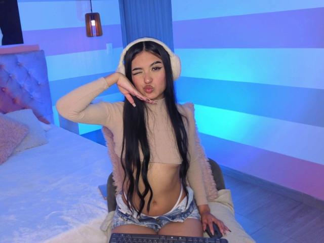 AmySaens met lang zwart haar, knipogend, gekleed in een beige topje, een witte hoed en denim shorts, zittend op een bed met haar tong uit, een toetsenbord voor zich..