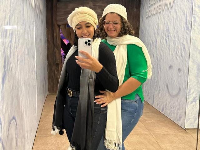 Marinomade with curly brown hair, green shirt, and white scarf stands behind Marinomade in black top and beige hat, taking selfie with white iPhone, both smiling.