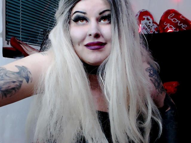Psychobitch7-hot with long, platinum blonde hair, dark purple lipstick, and tattoos on her arms, smiling at the camera, wearing a black top, with visible eyeliner and nose ring.