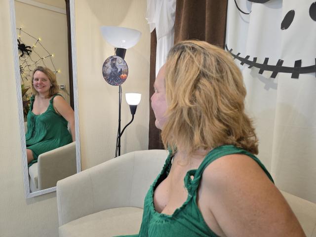 VestaWoody with blonde, wavy hair, green dress, showing small boobs, sitting in front of mirror, smiling, reflection visible, lamp and wall art in background.