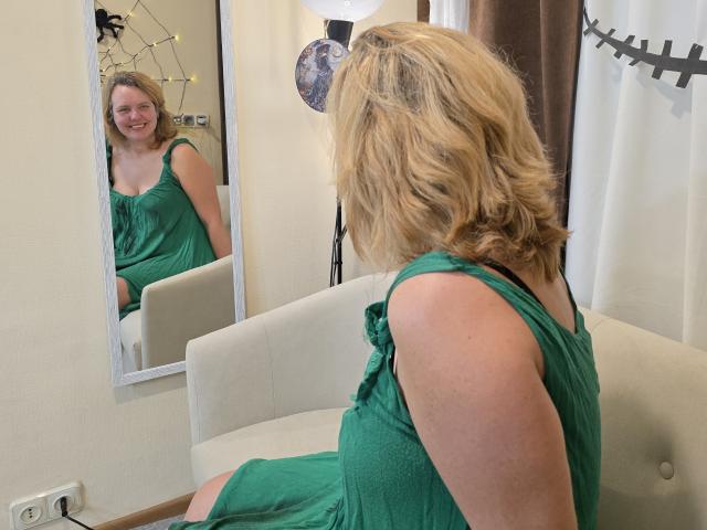 VestaWoody with blonde wavy hair, green dress, sitting on white sofa, facing mirror, smiling at reflection, showing her back and arms, with her tits slightly visible.