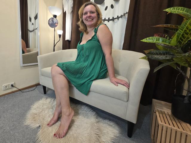 VestaWoody with pale skin, short blonde hair, and small tits, wearing a green dress, sits on a white sofa, smiling, barefoot on a fluffy rug, with a plant nearby.