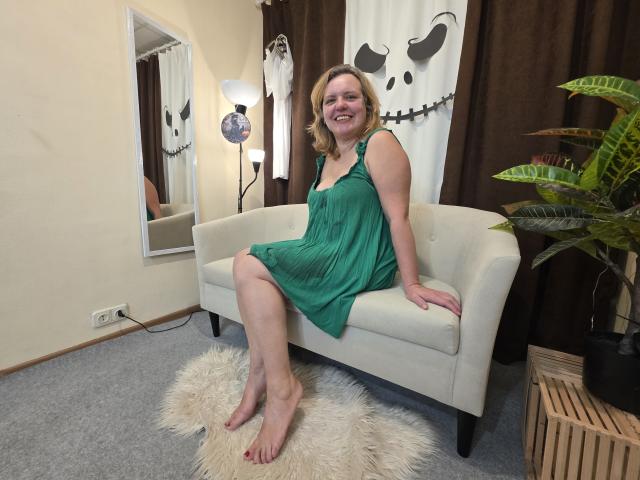 VestaWoody, barefoot with red toenails, sits on a white couch in a green dress, smiling, legs crossed, on a fluffy rug, with a potted plant to the right.