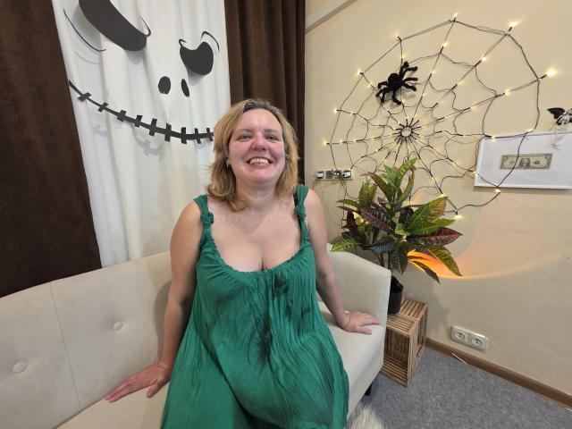 VestaWoody with blonde hair, green dress, and visible boobs, smiling, sitting on a white couch, background includes spider web decoration and plant.