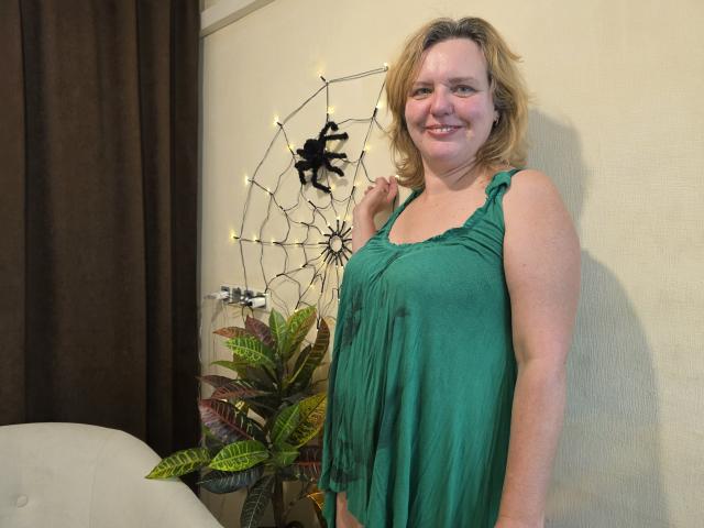 VestaWoody, a chubby woman with blonde hair, green dress, and small tits, stands smiling, holding her hair, next to a dreamcatcher and plant, in a room.