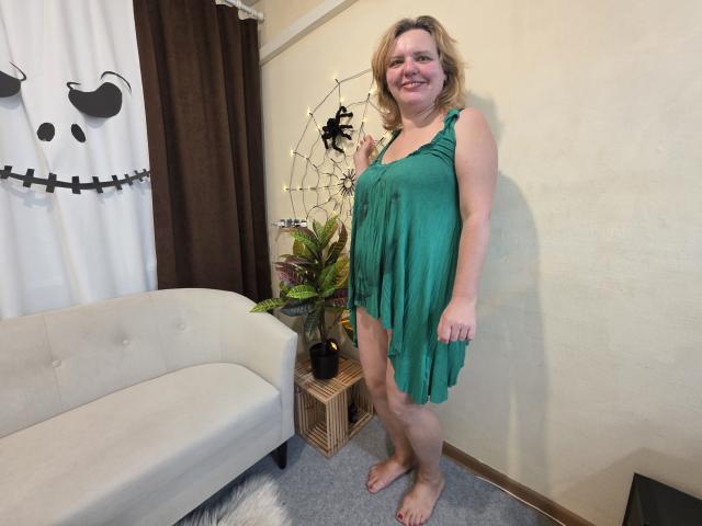 VestaWoody, a chubby woman with blonde hair, wearing a green dress, stands barefoot, smiling, with visible pussy through the dress, in a room with a white sofa.