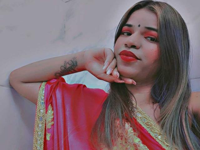 KairaDayamai with long brown hair, red lipstick, and a red sari with gold embroidery, resting her chin on her hand, showing a tattoo on her forearm.