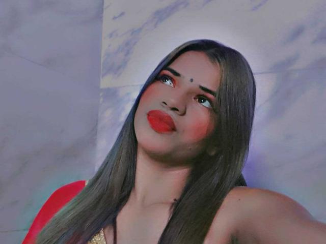 KairaDayamai with long dark hair, red lipstick, and red eye makeup, looking up with a playful expression, showing her bare shoulders and a gold top.