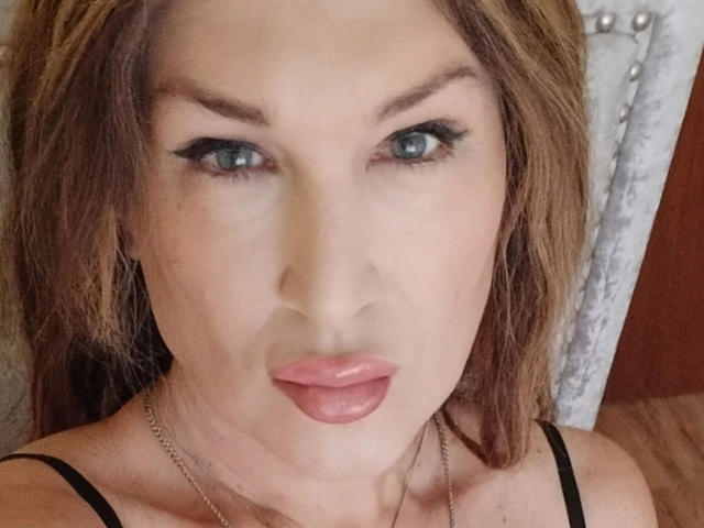 Tsnovaleex-hot has blue eyes, brown hair, black eyeliner, and pink lips. She's wearing a black strap and a silver chain. Her face is centered in the photo.