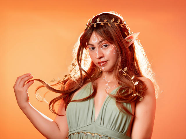 JuliettaHott with pointy elf ears, wearing a green dress, gold headpiece, and white face paint, holding her brown hair against an orange background. Boobs, ass, and tits are not visible.