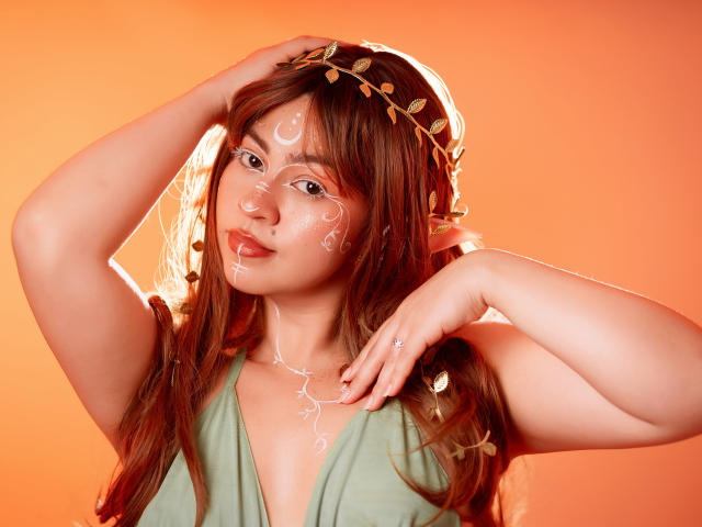 JuliettaHott with red hair, green dress, white face paint, leaf headband, hand in hair, orange background, shiny skin, cute, sexy, small nose ring, relaxed expression.