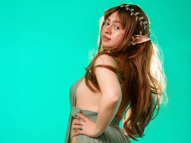 JuliettaHott with long brown hair, elf ears, white facial markings, and a green dress showing her back and sideboob, looking over her shoulder against a solid teal background.