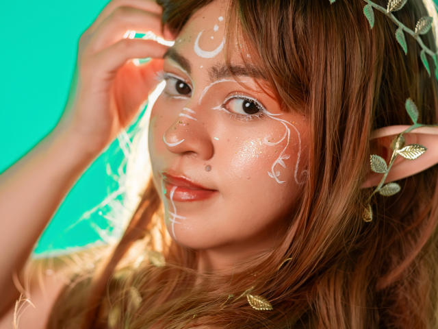 JuliettaHott with white face paint, pointed elf ears, and brown hair, has a cute nose piercing and shiny skin, against a teal background, looking slightly to the side.