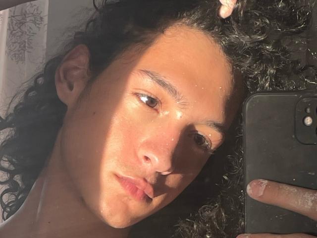 DiegoPhoenix with curly black hair, tan skin, and brown eyes, holding a phone, sunlight on face, slightly parted lips, finger on phone's side, casual selfie.