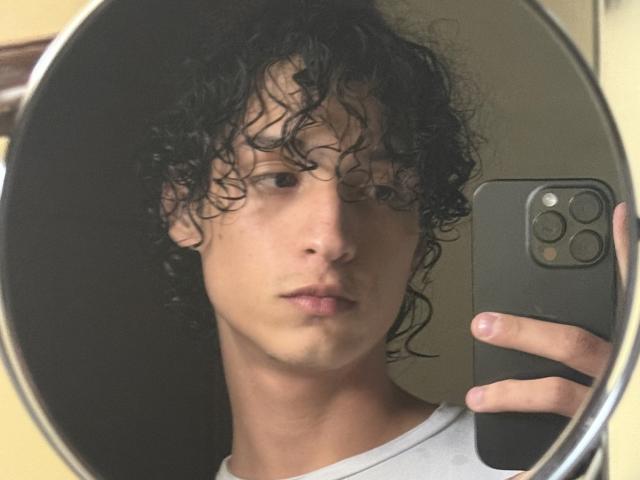 DiegoPhoenix with curly black hair, holding a phone, taking a mirror selfie, wearing a white shirt, looking serious, in a dimly lit room. Mirror reflection, iPhone in hand.