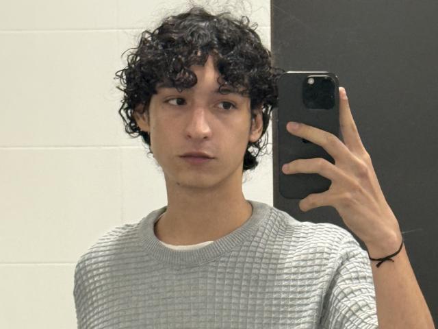 DiegoPhoenix with curly black hair, wearing a grey textured sweater, holding a black phone, taking a mirror selfie, looking slightly bored, wearing a black bracelet on his right wrist.
