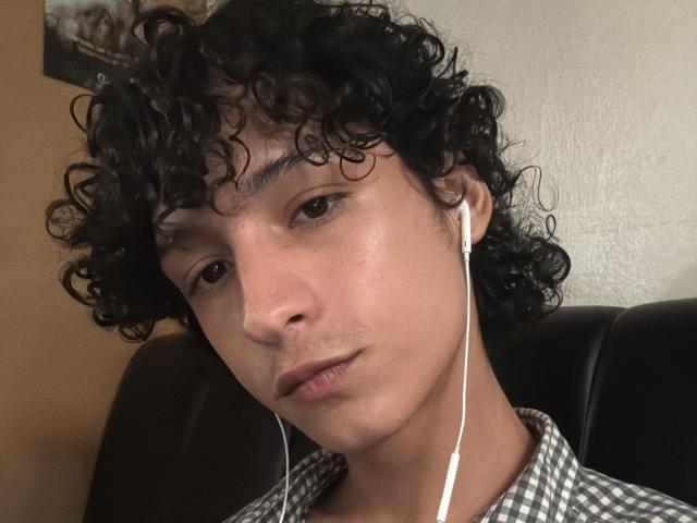 DiegoPhoenix with curly black hair, light brown skin, and white earbuds, wearing a checkered shirt, has a slight mustache and a relaxed, slightly smug expression.