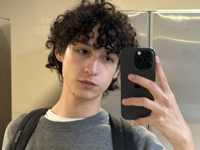 DiegoPhoenix with curly black hair, pale skin, and a neutral expression, holding a black phone, wearing a gray shirt, and a black backpack. Boobs not visible. Ass not visible.