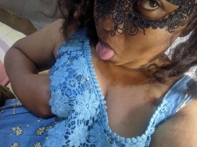 Leylahwilliam-hot with dark curly hair, wearing a black lace mask, sticking out her tongue, and showing her blue lace-up boobs with a patterned dress.