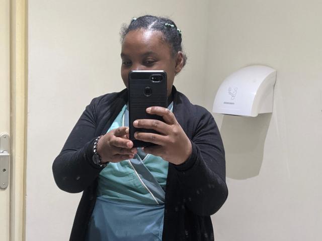 Leylahwilliam-hot, dark-skinned, with small, round tits, taking a selfie in a bathroom, wearing a black cardigan over a teal shirt, braided hair with green clips.