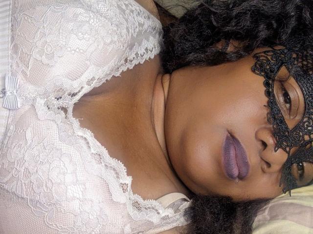 Leylahwilliam-hot with dark curly hair, wearing a lace mask and white lace top, showing her purple lipstick and brown skin. Boobs visible through lace. Eyebrow raised slightly.