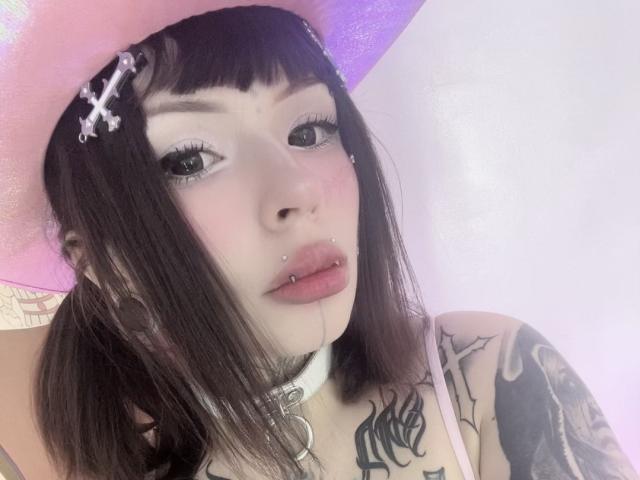 StarWitch with pink hat, black hair, tattoos, white choker, pierced nose, and lip, wearing makeup, looking at camera. Boobs partially visible, pink hat with white cross hairpins.