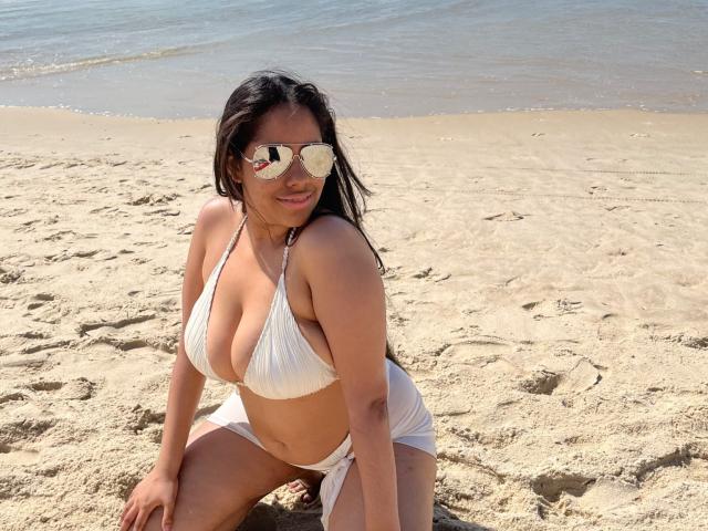 SiennaTaylor with large tits in a white bikini, wearing reflective sunglasses, kneeling on sandy beach, looking at camera, sunlight on tan skin, black hair, ocean in background.
