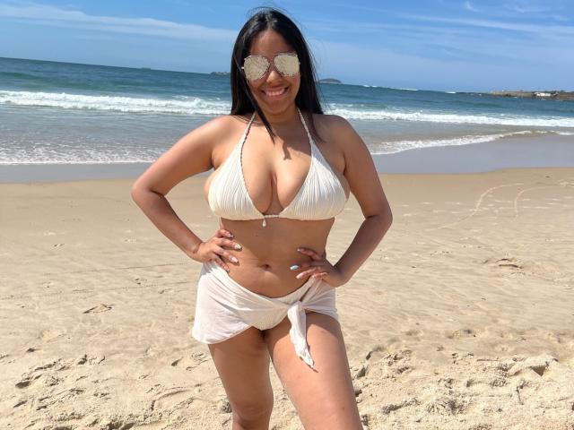 SiennaTaylor with big boobs, wearing a white bikini top and tied white skirt, standing on a sandy beach, smiling, hands on hips, wearing reflective sunglasses, dark hair.