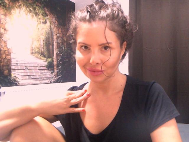 SofiaLiub has curly brown hair, wearing a black shirt, sitting with her hand near her neck, pouting with pink lips, and red nails, in a softly lit room.