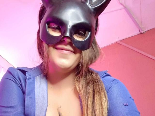 CleareRems with a black mask, smiling, wearing a blue shirt, brown hair in a ponytail, showing a small tattoo above her cleavage, pink light in the background.
