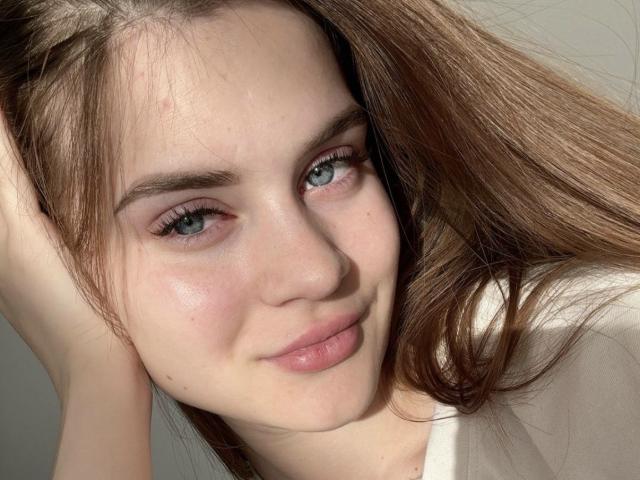 LuisaClover has light blue eyes, fair skin, and long brown hair. She's smiling slightly with her hand in her hair, wearing a white shirt. Her ass is not visible.