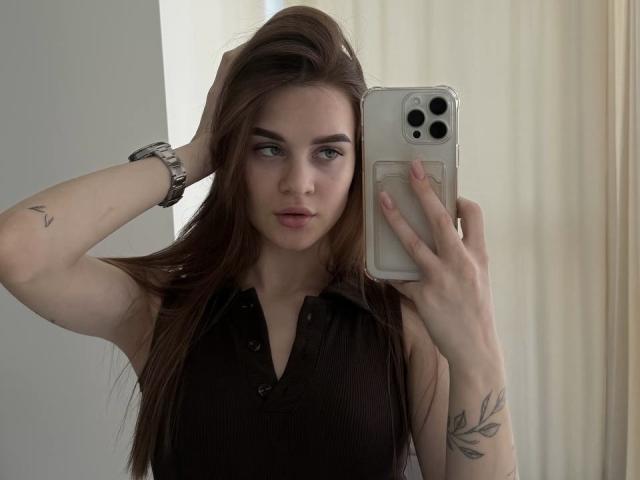 LuisaClover with long brown hair, wearing a black sleeveless shirt, holding a phone, tattoo on forearm, wristwatch, serious expression, looking into mirror, visible breasts.