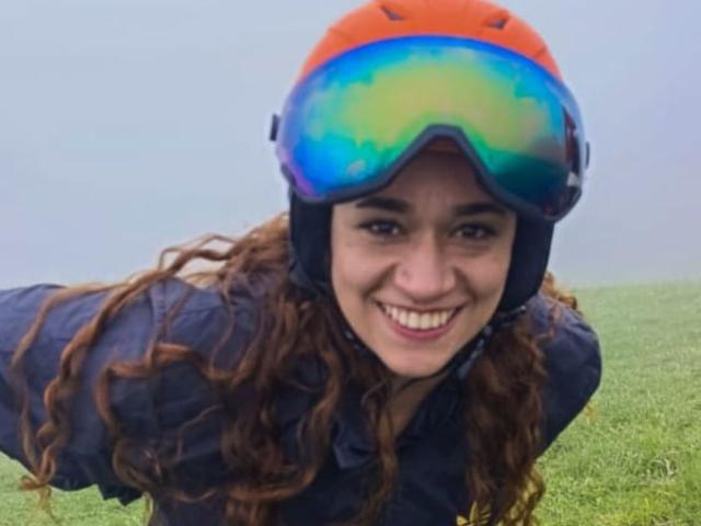 ValeryHilton with curly brown hair, smiling, wearing an orange helmet and blue-green ski goggles, crouched in green grass, wearing a dark blue jacket.
