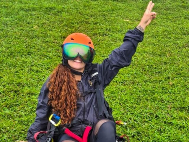 ValeryHilton with curly red hair, wearing an orange helmet, black jacket, and reflective goggles, raising her right arm, sitting on grass with climbing gear.