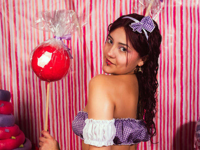 EmilyLorenz with curly brown hair, wearing a purple checkered off-shoulder top, holding a red candy lollipop, looking over her shoulder with a playful smile, pink and white striped background.
