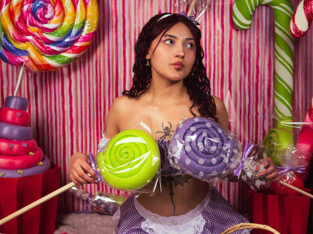 EmilyLorenz with braided hair, holding huge green and purple swirl lollipops, wearing a checkered dress, her tits covered in plastic, and a spider tattoo on her chest.