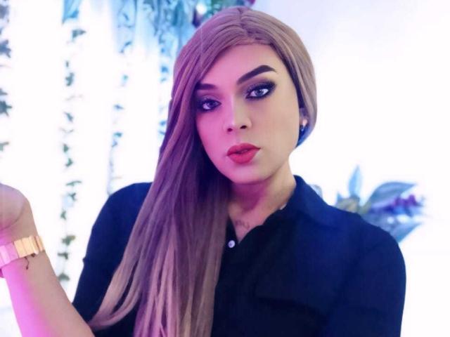 KarolElegance with long brown hair, black shirt, red lipstick, and gold watch, looking at camera with a slight pout. She's got bold eyebrows and dark, dramatic eye makeup.