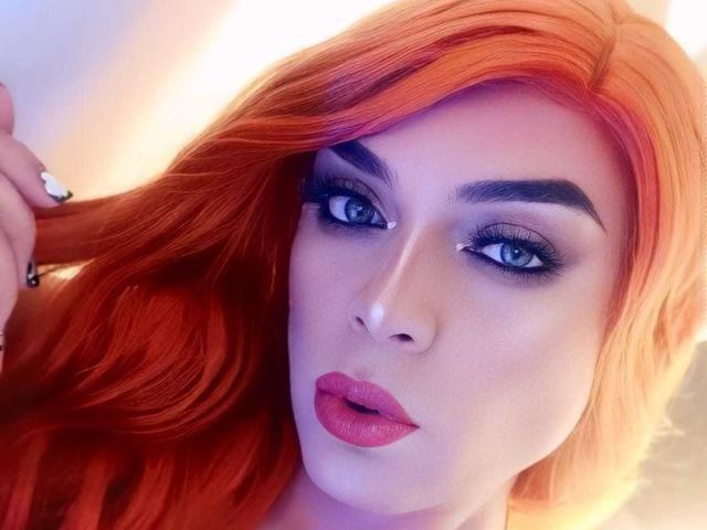 KarolElegance has bright orange hair, blue eyes, and red lipstick. Her eyebrows are dark and she's touching her hair with a ringed finger. Her face is slightly pouty.